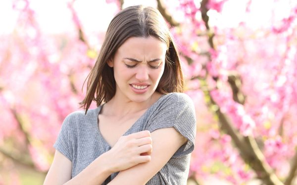 Blogbild – eine Person kratzt sich im Freien vor blühenden rosa Zweigen am Arm, etwa nach einem Mückenstich