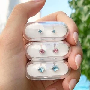 Hand hält drei Blisterpackungen mit Blomdahl Ohrsteckern aus medizinischem Kunststoff – blaue, rosa und hellblaue blütenförmige Steine